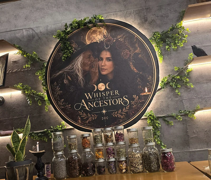 Jars of dried petals and herbs on the studio workbench beneath the elder-and-Laiany portrait, surrounded by trailing greenery.