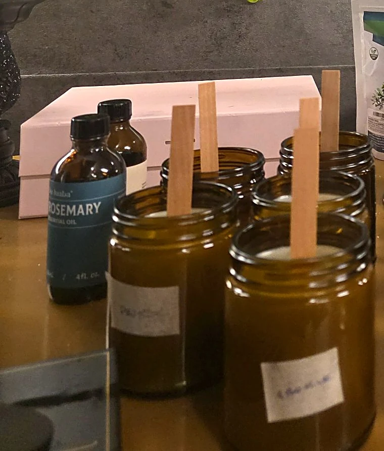 Amber candle jars with upright wooden wicks on Laiany's workbench, beside bottles and candle-making supplies.